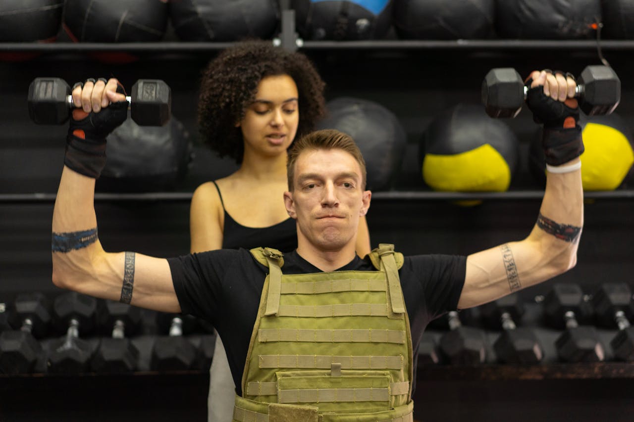 services-01 A man in adaptive training gear lifts dumbbells with support from a trainer in a gym.